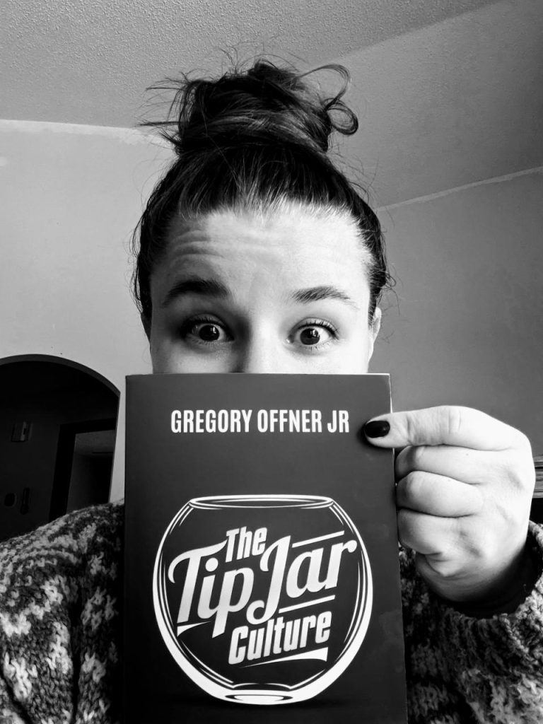 A person posing with a copy of the book, "The Tip Jar Culture."